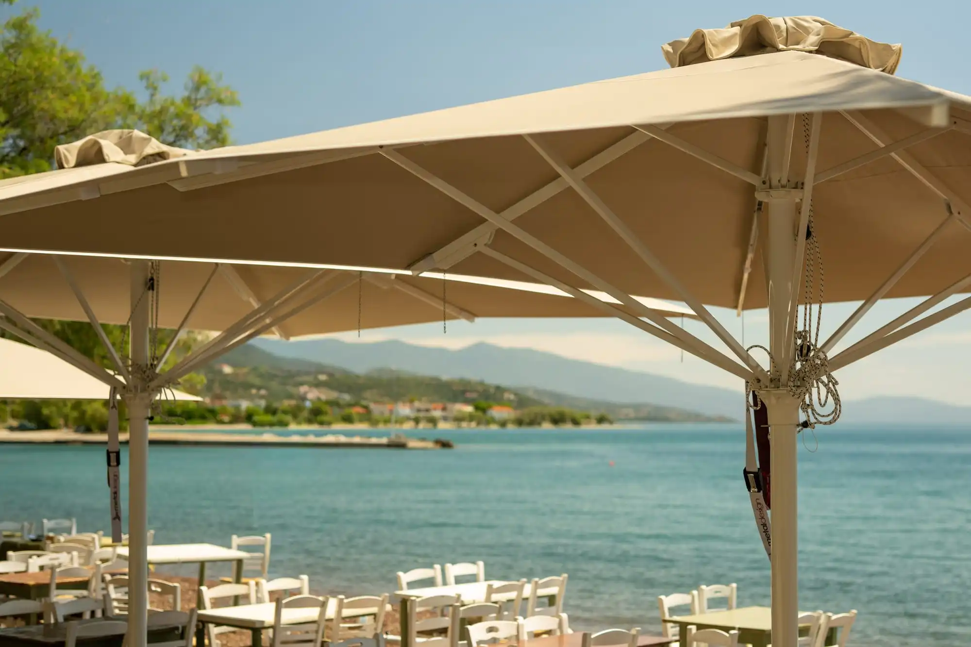 Δύο κρεμ ομπρέλες Metal Design πάνω από τραπέζια και καρέκλες δίπλα στη θάλασσα