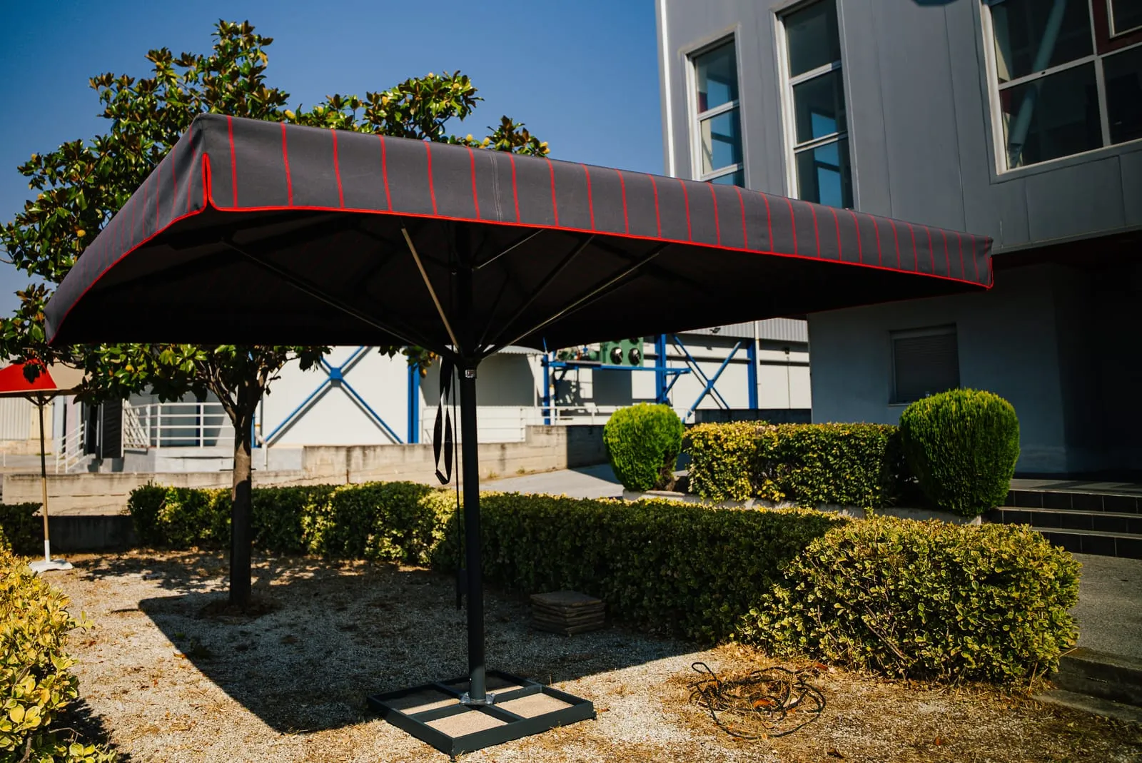 Μεγάλη ομπρέλα Metal Design με σκούρο γκρι ύφασμα και κόκκινες ρίγες μπροστά στο κτίριο της εταιρείας στον Βόλο